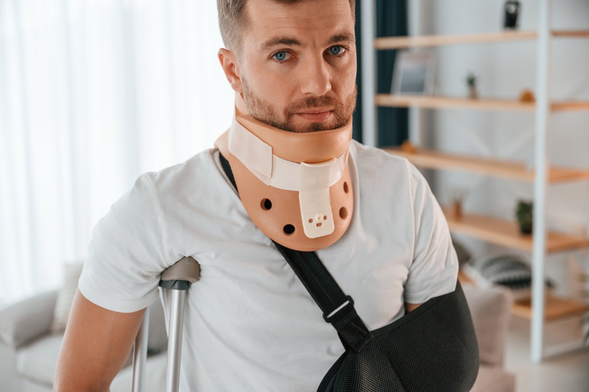 Injured man with neck brace and arm sling using crutches at home