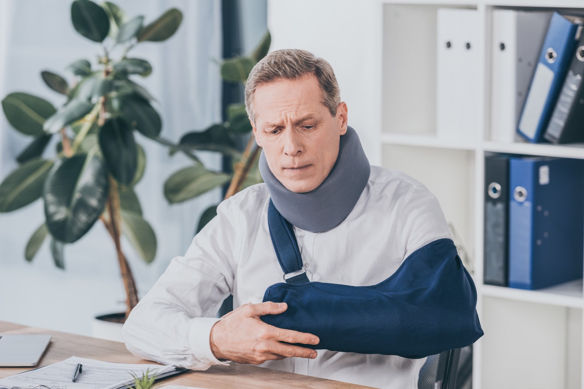 Man with neck brace and arm sling holding his injury in office